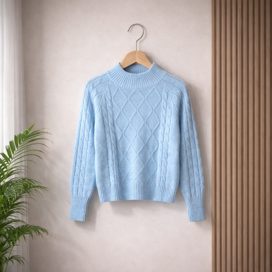Light blue cable‑knit sweater with diamond and sleeve patterns, hanging on a wooden hanger against a beige wall with a potted plant nearby.