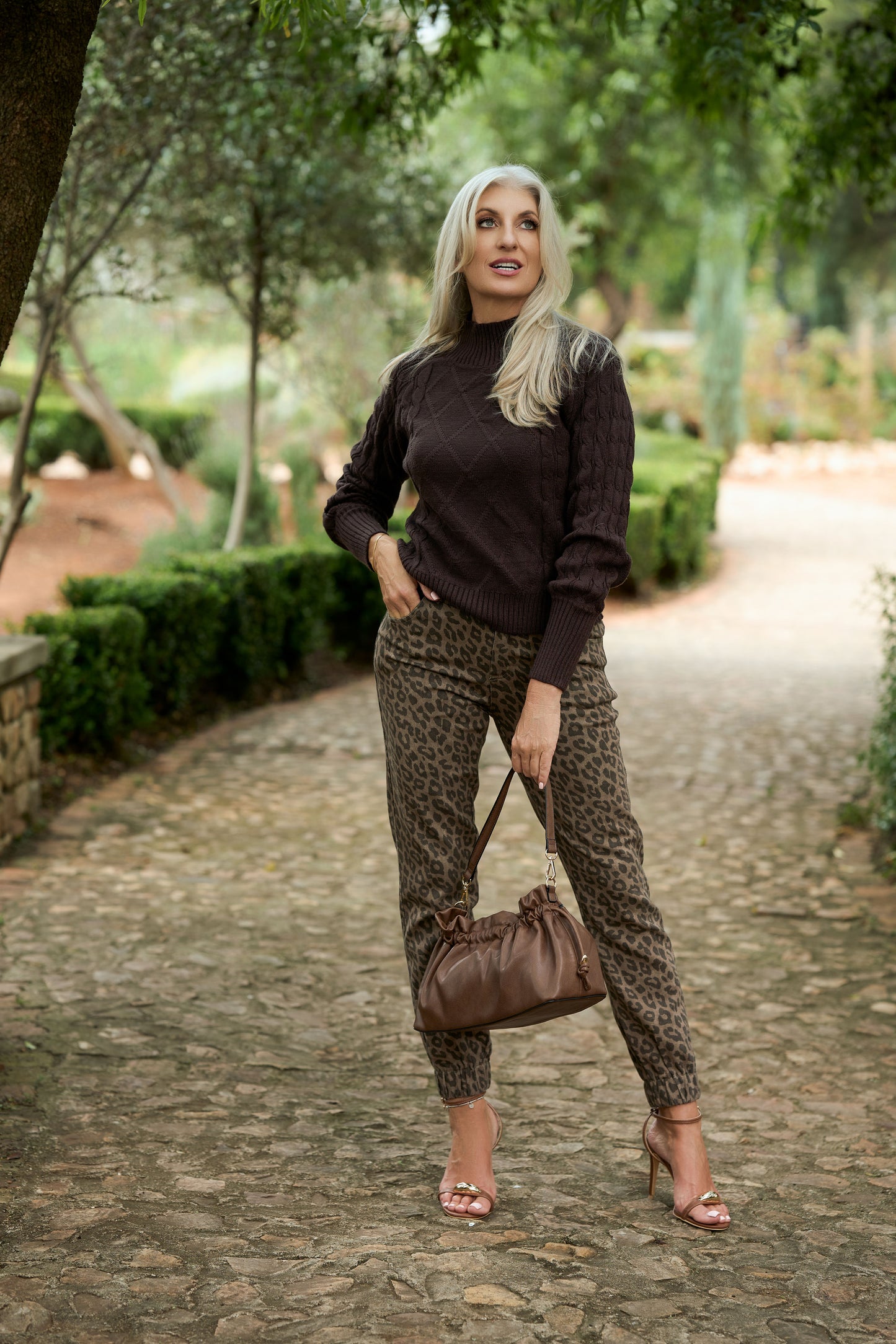Person wearing a dark brown cable‑knit sweater with leopard‑print pants and sandals, holding a brown handbag while standing on a cobblestone path surrounded by greenery