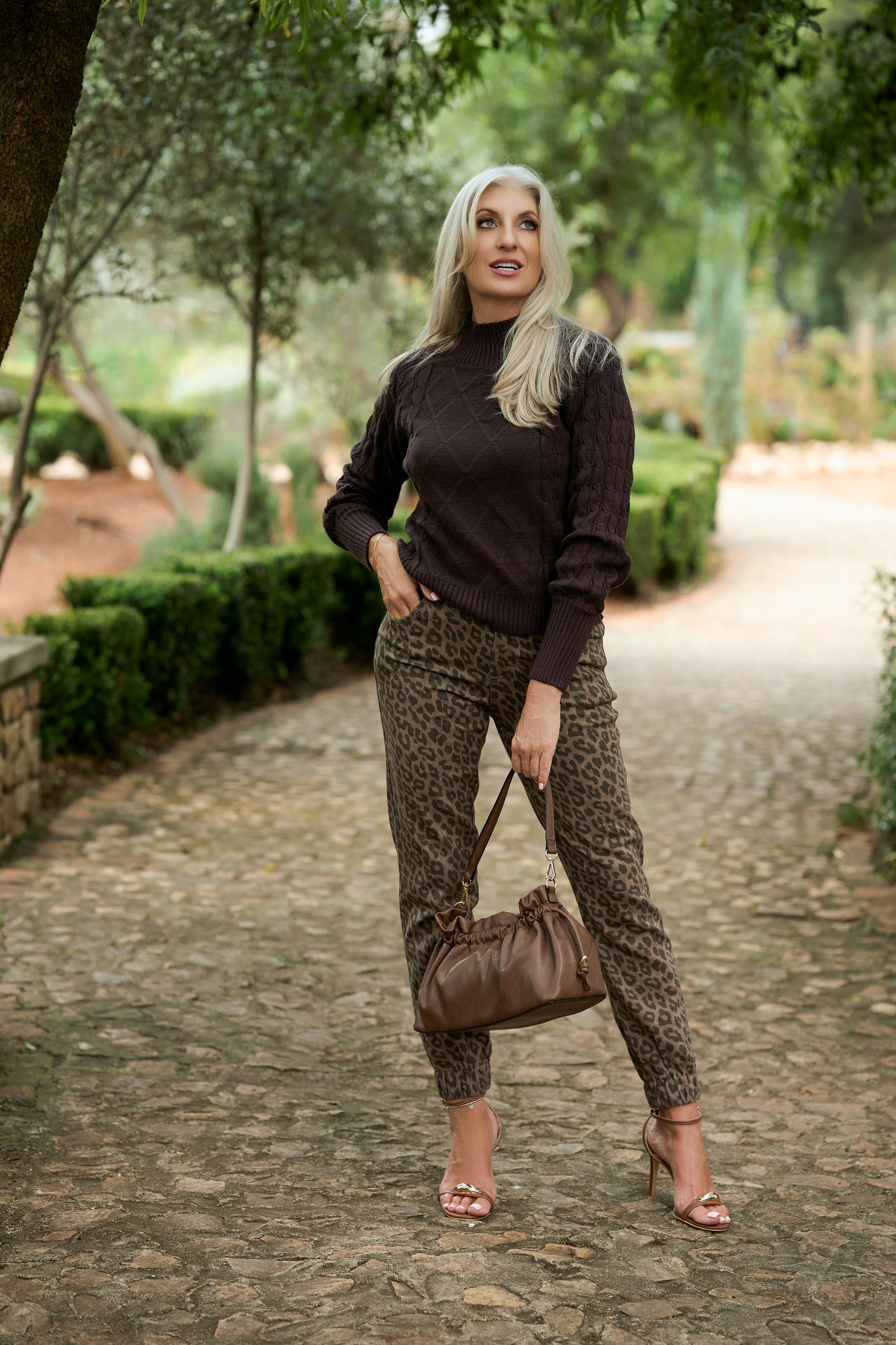 Person wearing a dark brown cable‑knit sweater with leopard‑print pants and sandals, holding a brown handbag while standing on a cobblestone path surrounded by greenery