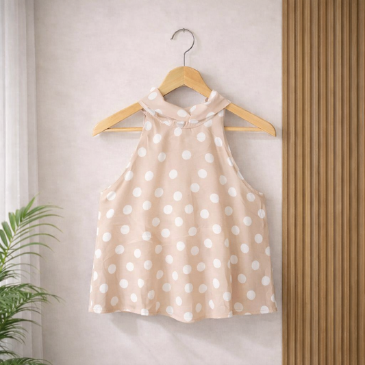 Blush Nude sleeveless halter blouse with white polka dots hanging on a wooden hanger against a neutral wall, with a plant and wooden slats in the background
