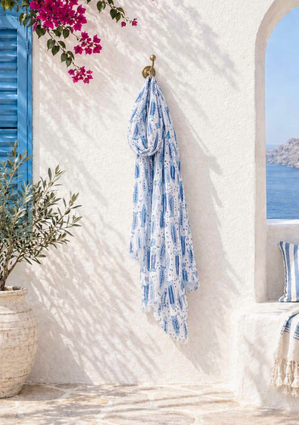 A blue and white patterned scarf or shawl hangs from a brass hook on a white stucco wall in a sunlit Mediterranean-style setting, with flowers, a potted olive tree, blue shutters, and a window view of the sea in the background.