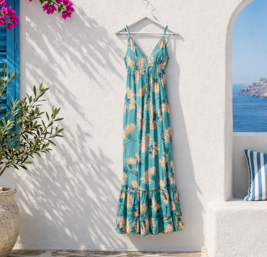 A teal floral maxi dress with peach and cream flower prints hangs from a white hanger against a sunlit Mediterranean-style white wall. The dress has thin straps, a V-neckline, and a ruffled hem, appearing slim and elegant