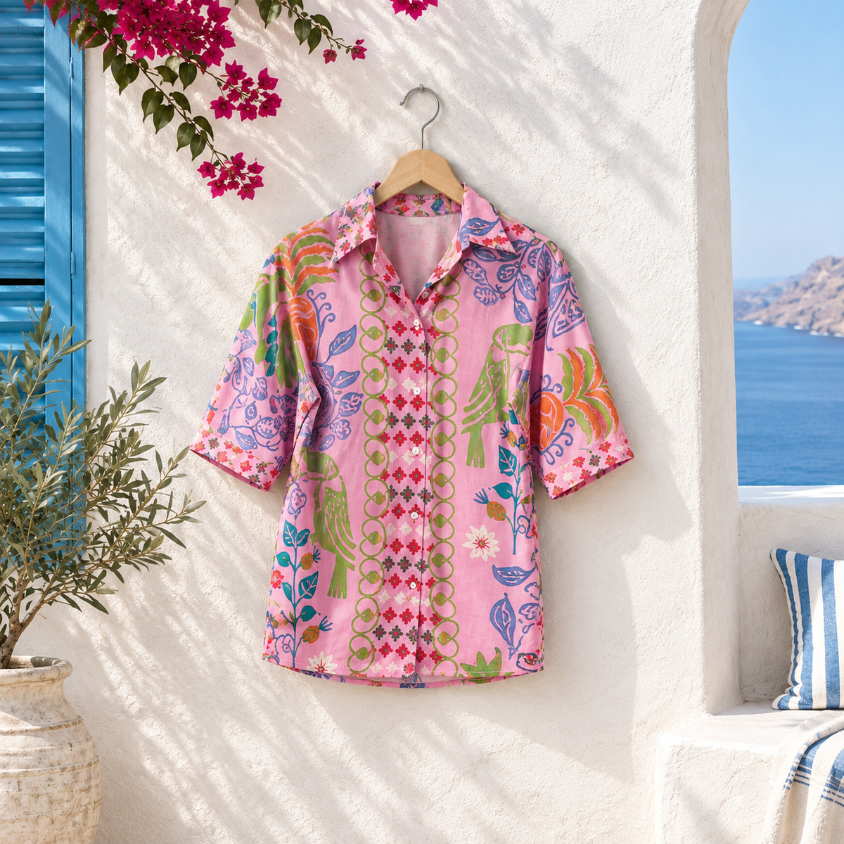 Pink patterned short-sleeve button-up shirt with colourful floral, paisley and geometric prints, displayed on a wooden hanger in a Greek-inspired setting
