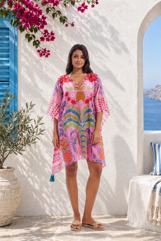 A woman modeling a vibrant pink kaftan dress with a multicolor tropical and floral print, standing in front of a white stucco wall with blue shutters and a view of the sea in the background.