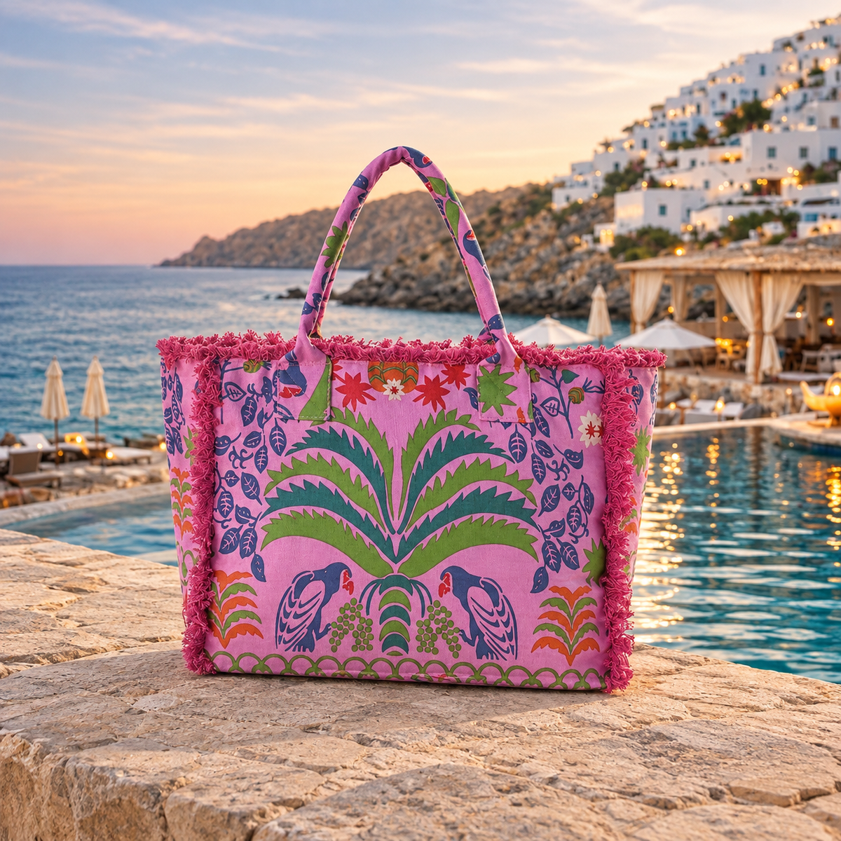 A pink tote bag with a colorful tropical print and pink fringe, set on a stone ledge next to a pool with a scenic Mediterranean coastal background at sunset.