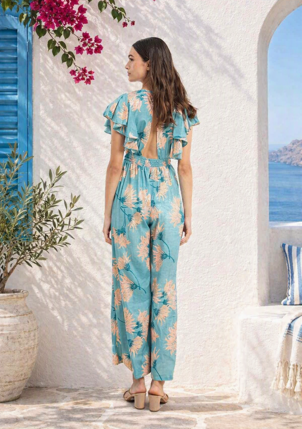 Floral jumpsuit with V‑neckline, short ruffled sleeves, and flowing skirt detail hanging on wooden hanger against Mediterranean white stucco wall, framed by pink bougainvillea, olive plant, and sea view.