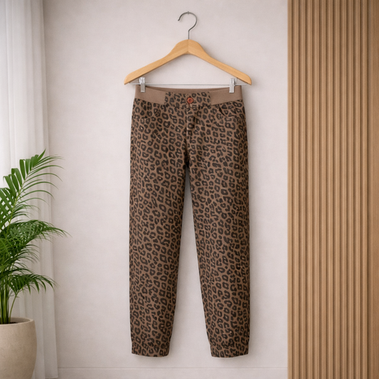 Leopard-print pants with a fitted waist and tapered legs hanging neatly from a light wooden hanger against a minimalist interior background featuring a potted green plant and vertical wooden slat panel