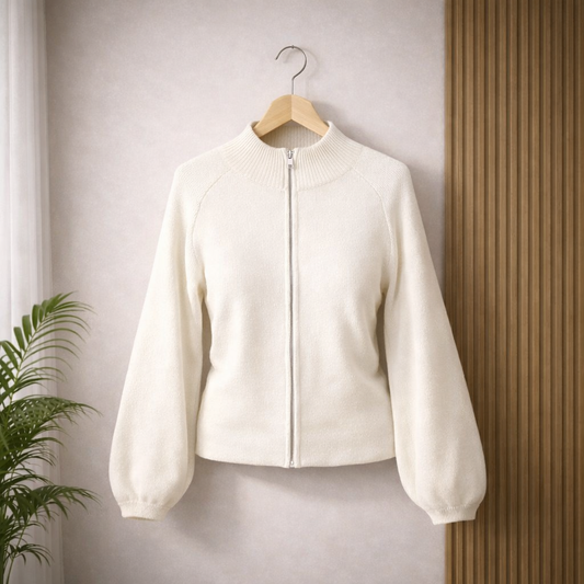 Off-white zip-up cardigan with a ribbed high collar and puffed sleeves, hanging on a wooden hanger against a neutral wall with subtle shadows and a green plant in the background