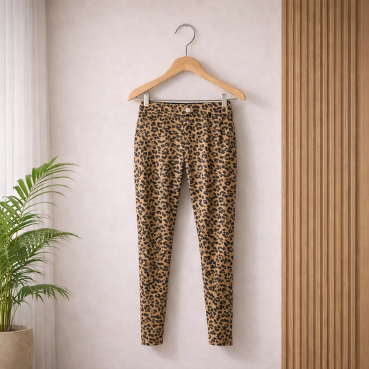 Leopard print pants hanging on a wooden hanger against a light wall with a plant in the corner.