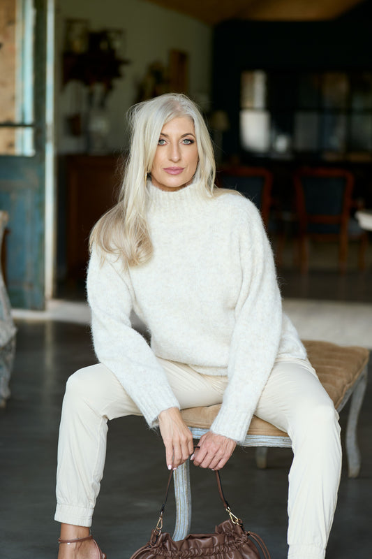Woman sitting on a bench wearing ZADE Collection cream-colored tailored joggers with a white knit turtleneck sweater and brown leather accessories.